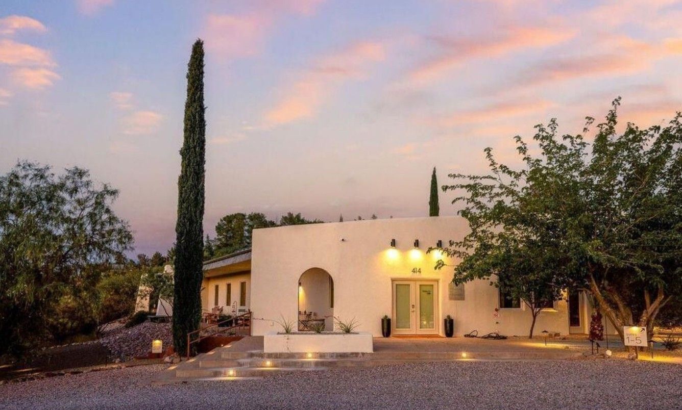 White stucco house with arched doorway, tall cypress tree, and gravel driveway at sunset.