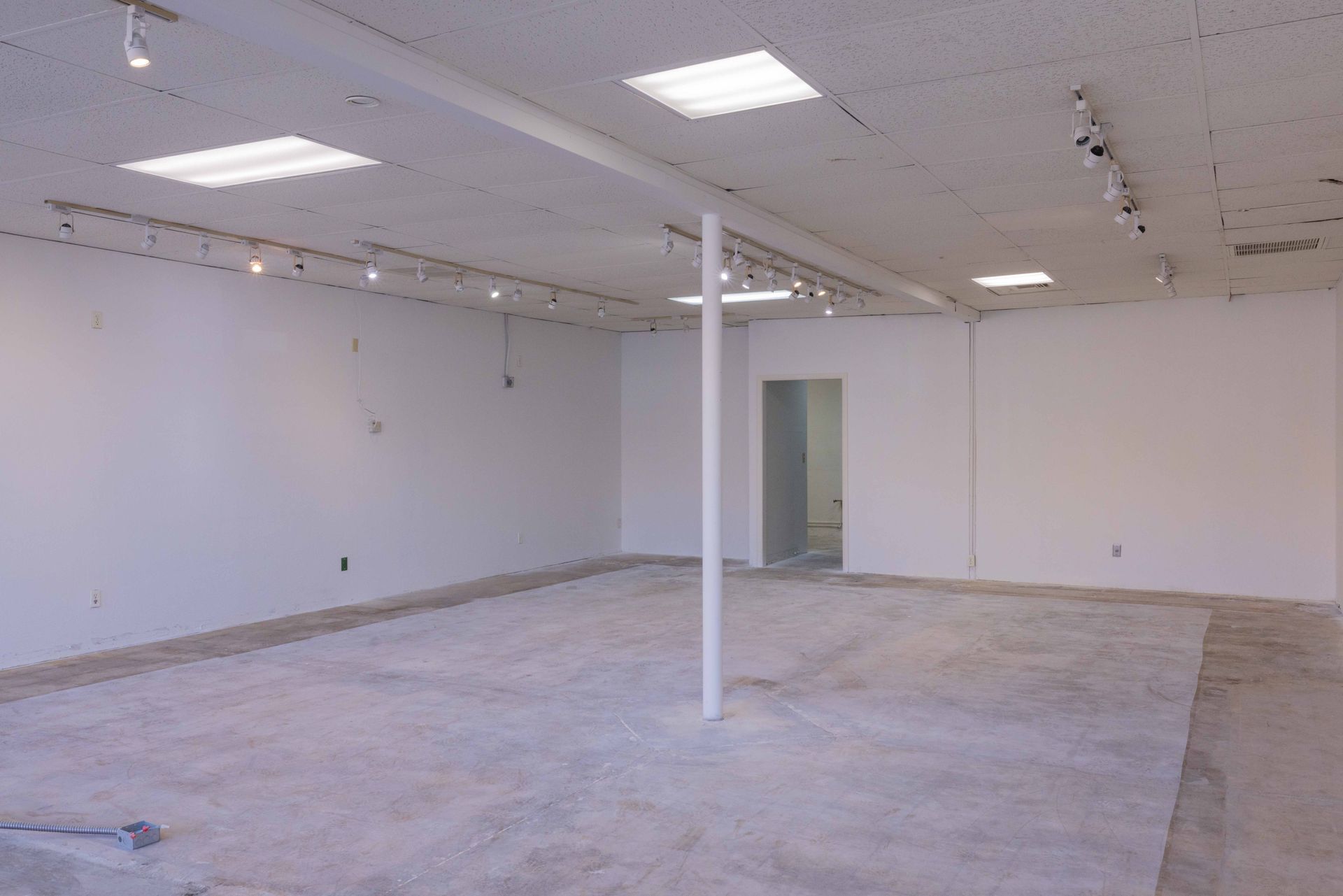 Empty, white-walled room with concrete floor and track lighting, a support pole, and a doorway.