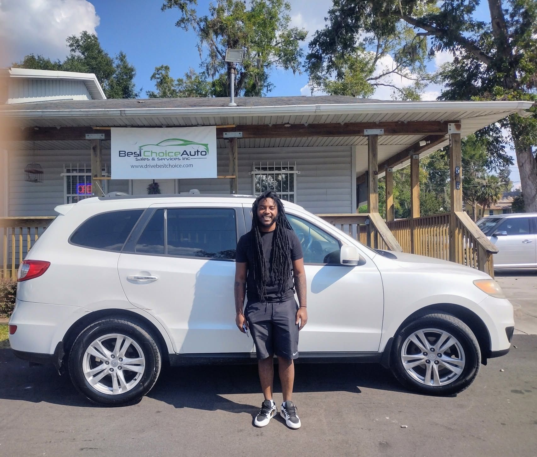 Person standing beside a white SUV in front of a small building on a sunny day