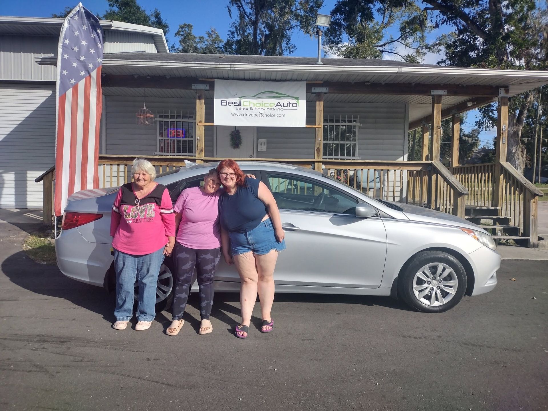 Three people stand beside a silver car in front of a house with an American flag.