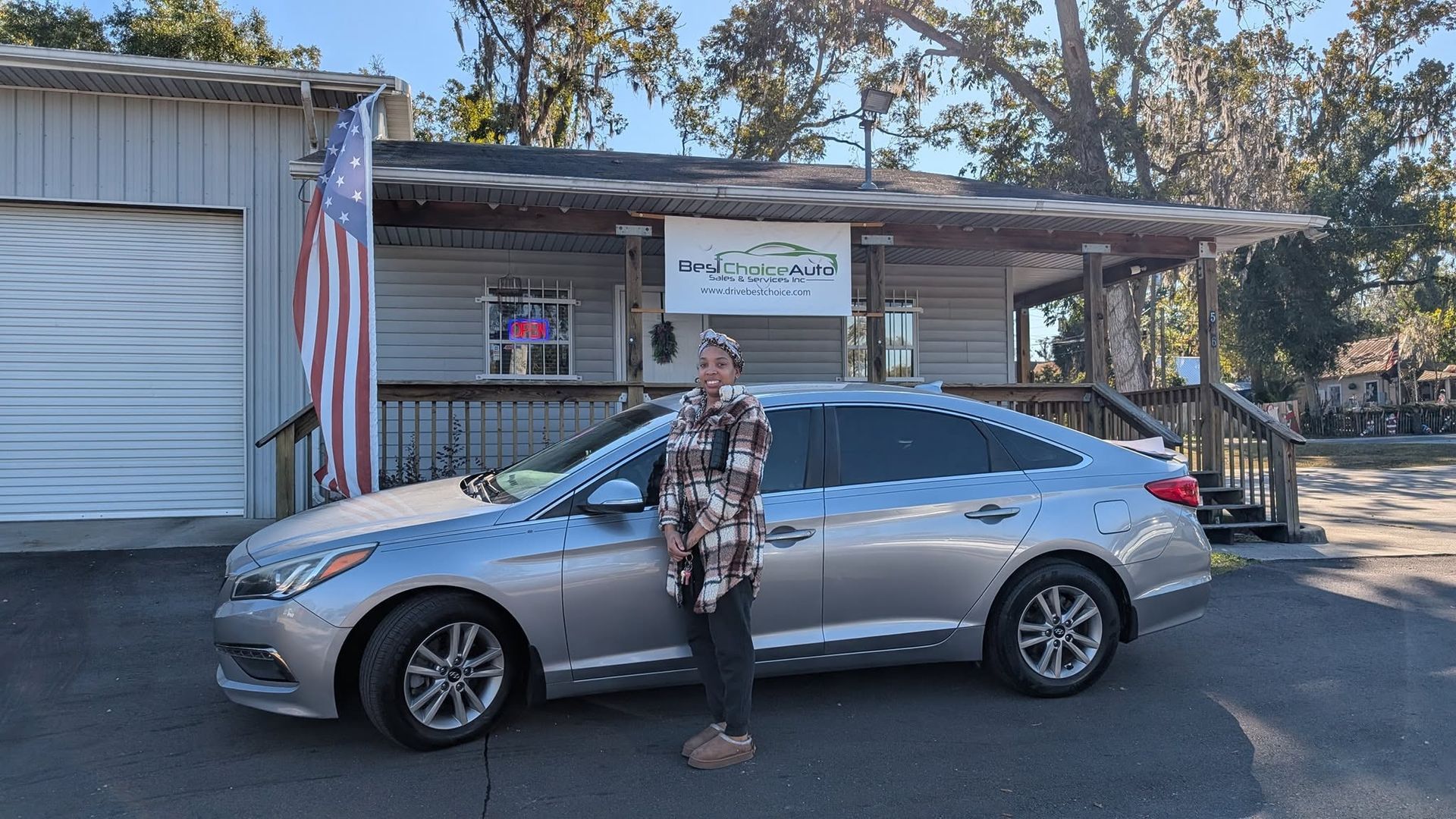 Person standing beside a silver sedan parked in front of a small building with an American flag.