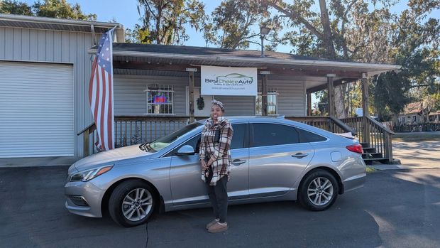 Person standing beside a silver sedan in front of a small storefront with an American flag.
