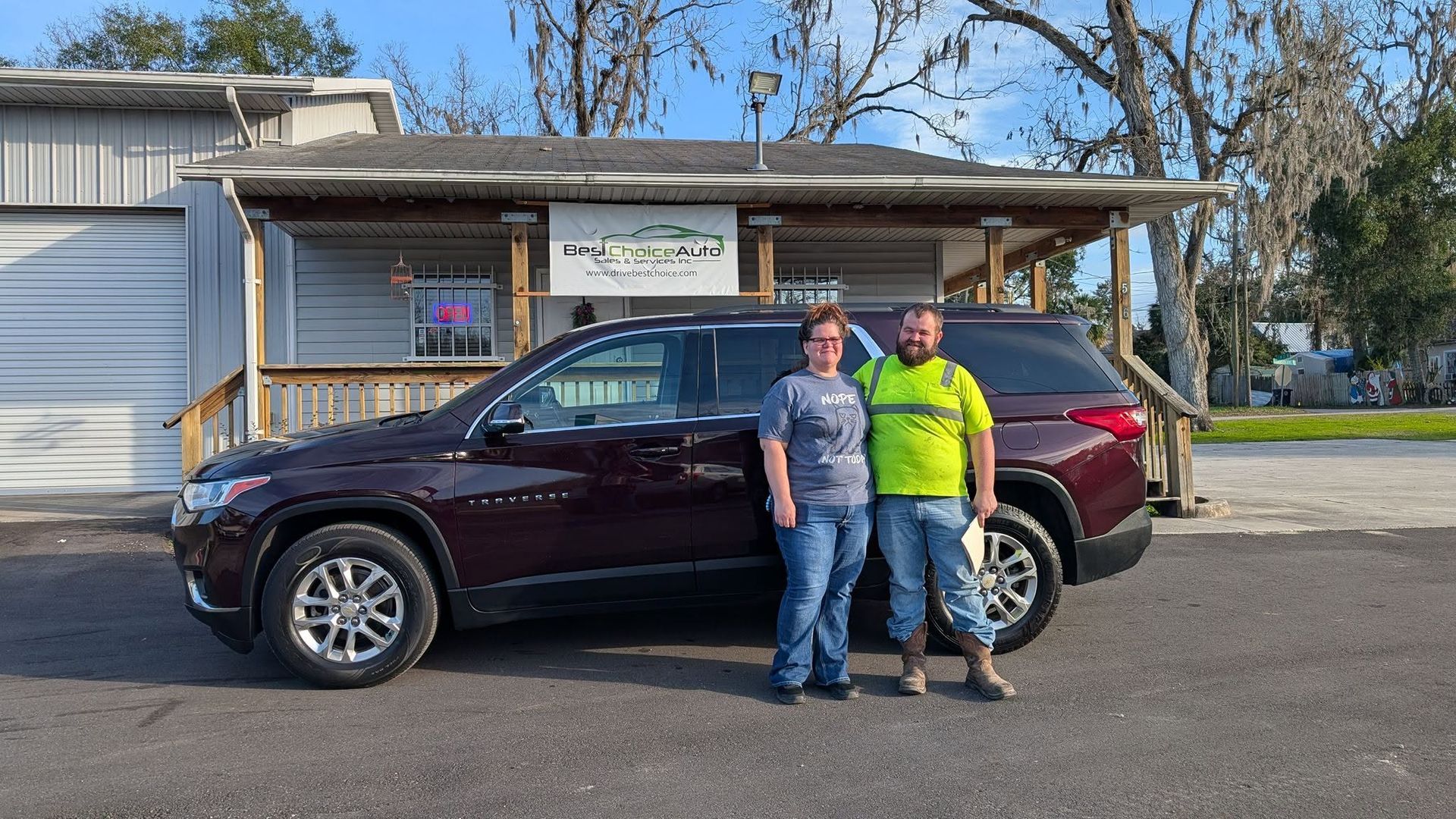 Two people standing beside a maroon SUV in front of a small building on a sunny day