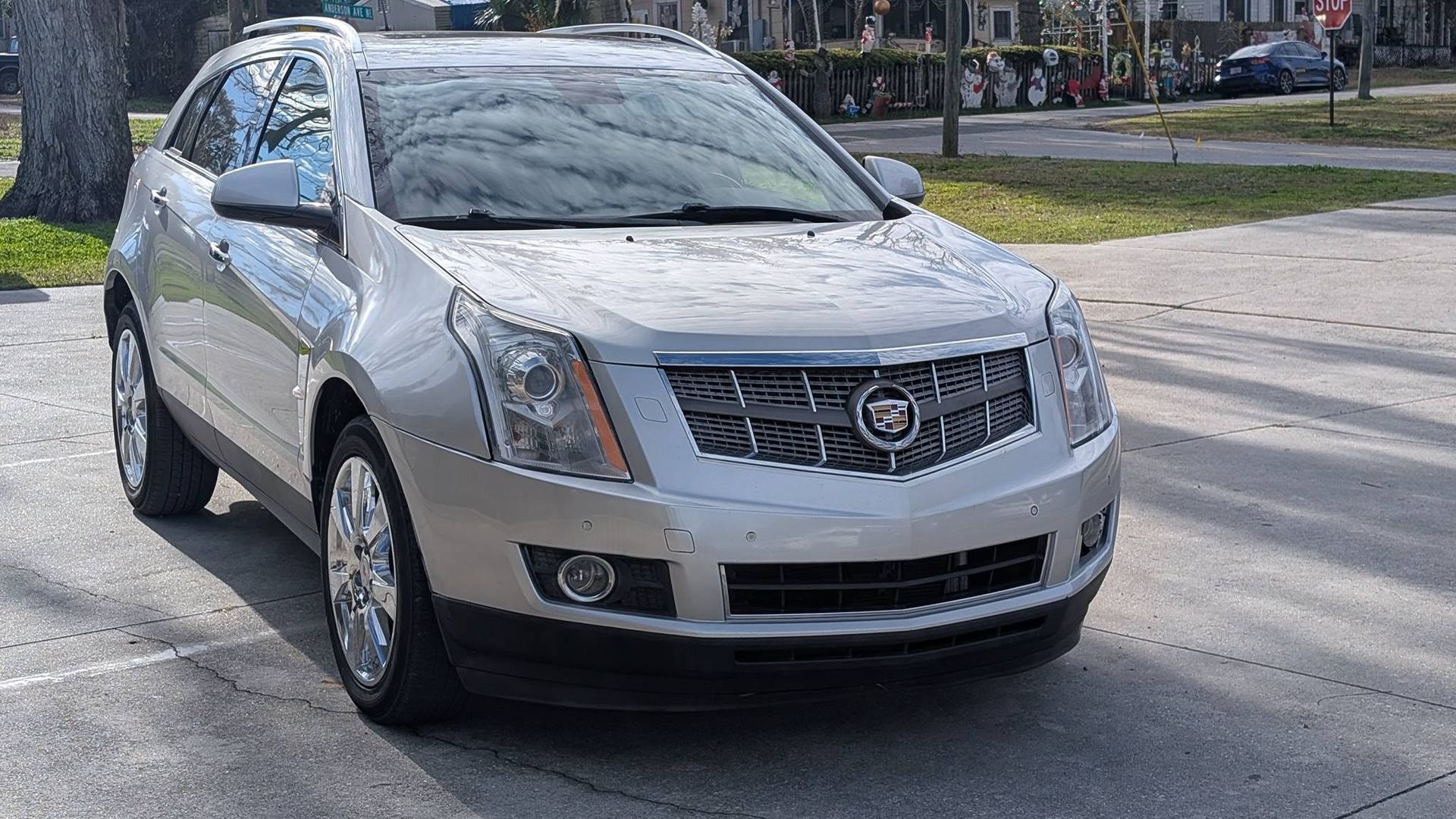 Silver Cadillac SUV parked on a sunny street, front-left view with city buildings in the background