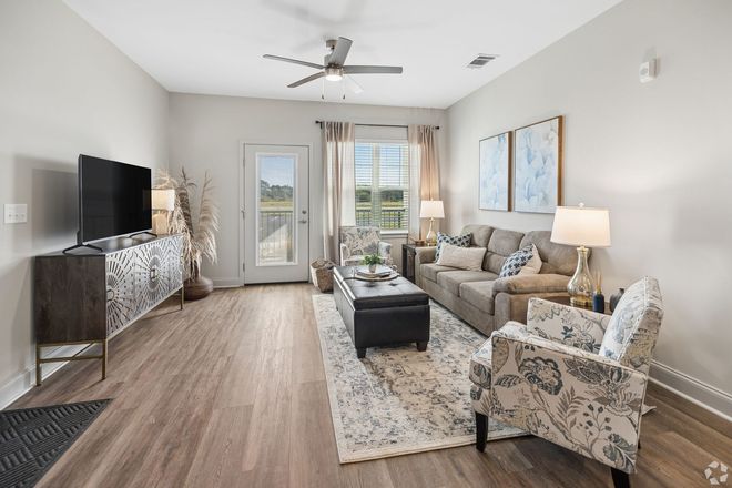 Bright modern living room with gray sofa, patterned chair, TV, coffee table, and large windows.