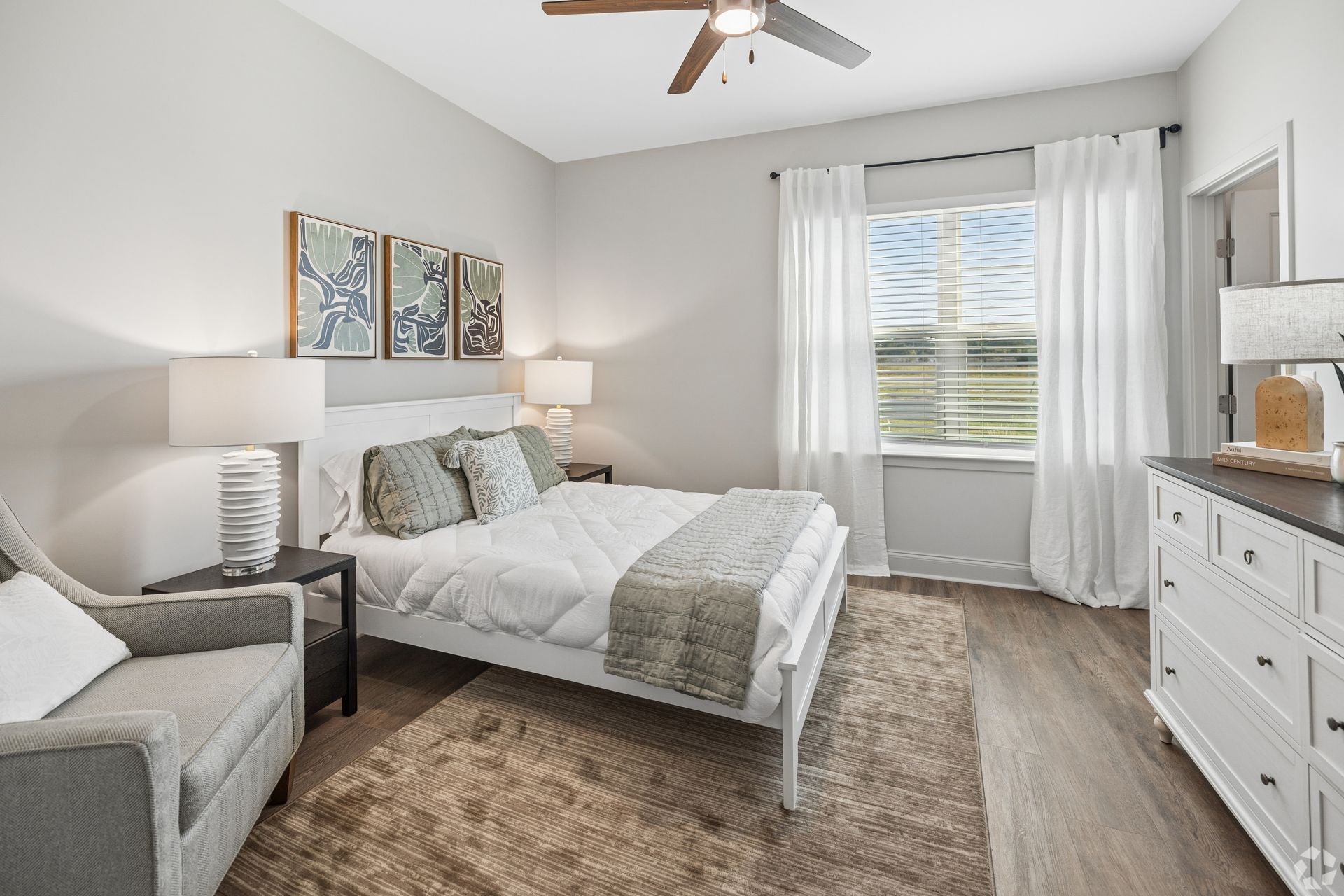 Bright bedroom with a bed, armchair, dresser, and window with white curtains