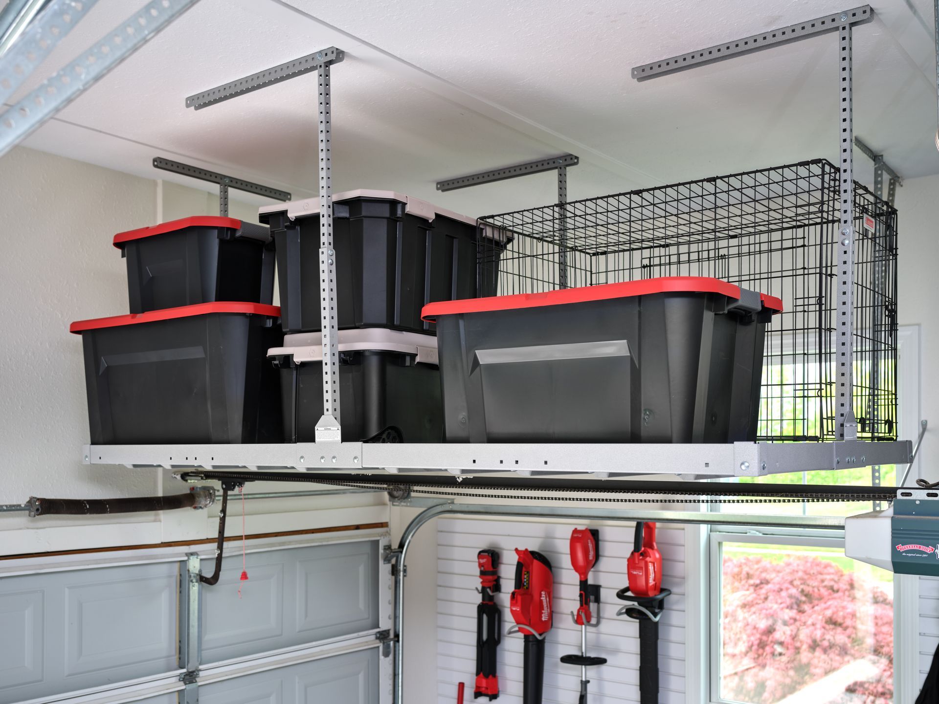 Garage ceiling storage shelves with black and red bins and hanging tools above closed cabinets