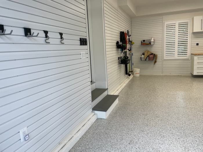 Empty garage with white slat walls, gray speckled floor, and shelves along the back wall.