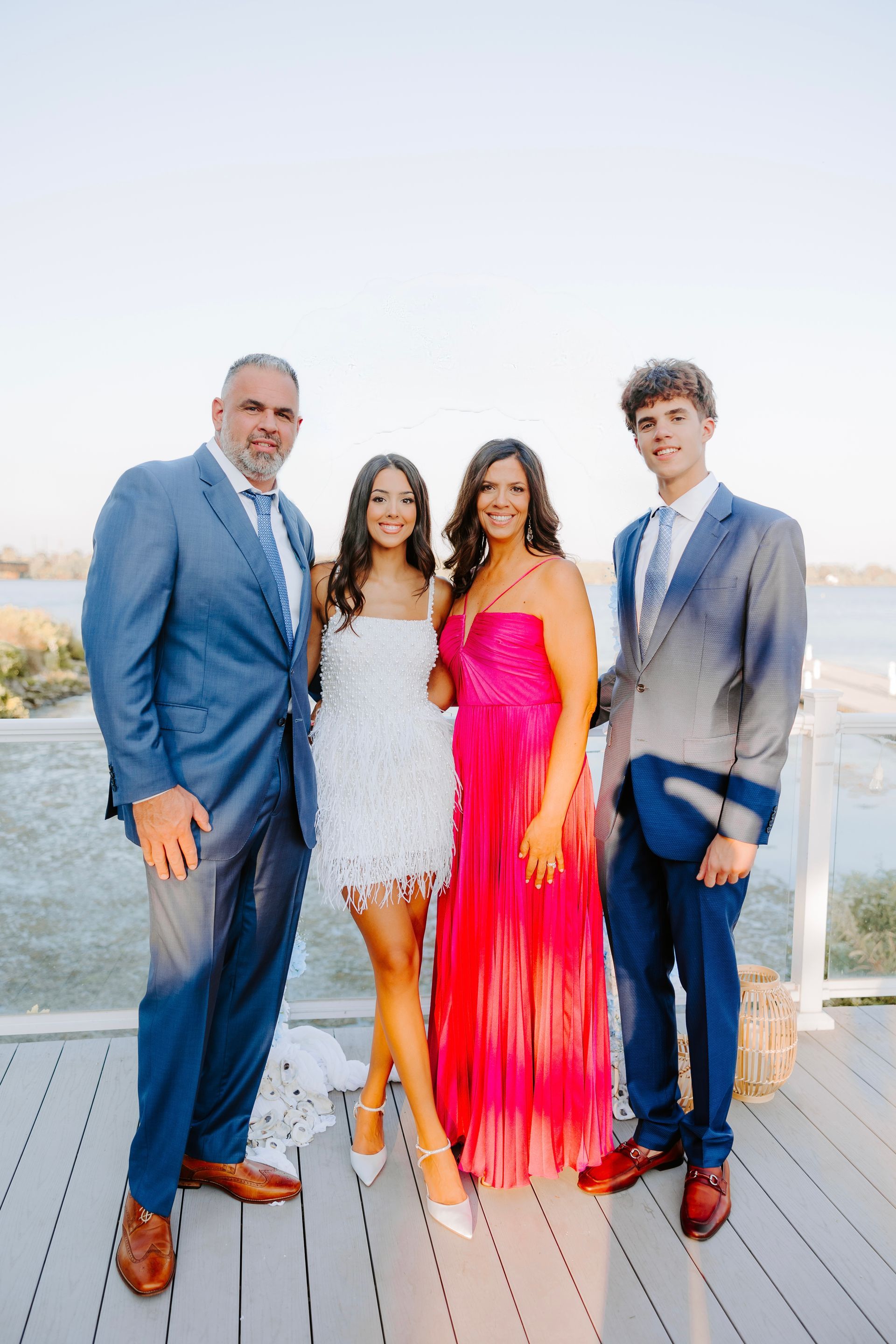 Four people posing outdoors on a dock, wearing formal outfits, with water in the background.