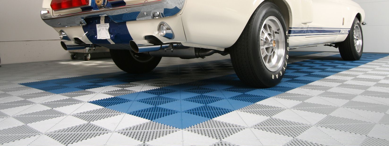 Rear view of a classic white car on a blue-and-gray checkered floor, showing chrome bumper and dual exhausts