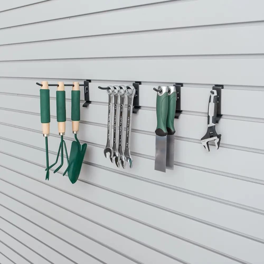 Garden tools hanging on black wall hooks against gray siding, including shovels, wrenches, and gloves.