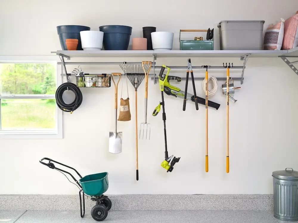 Garage wall with hanging garden tools, buckets, and a hose above a small wagon and trash can