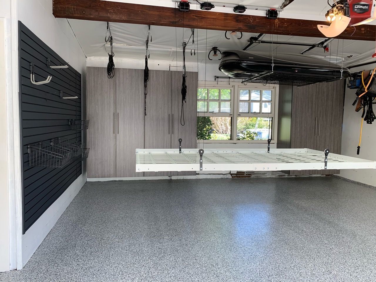 Empty garage with gray walls, hanging tracks, exposed beams, and a speckled floor near a window and workbench.