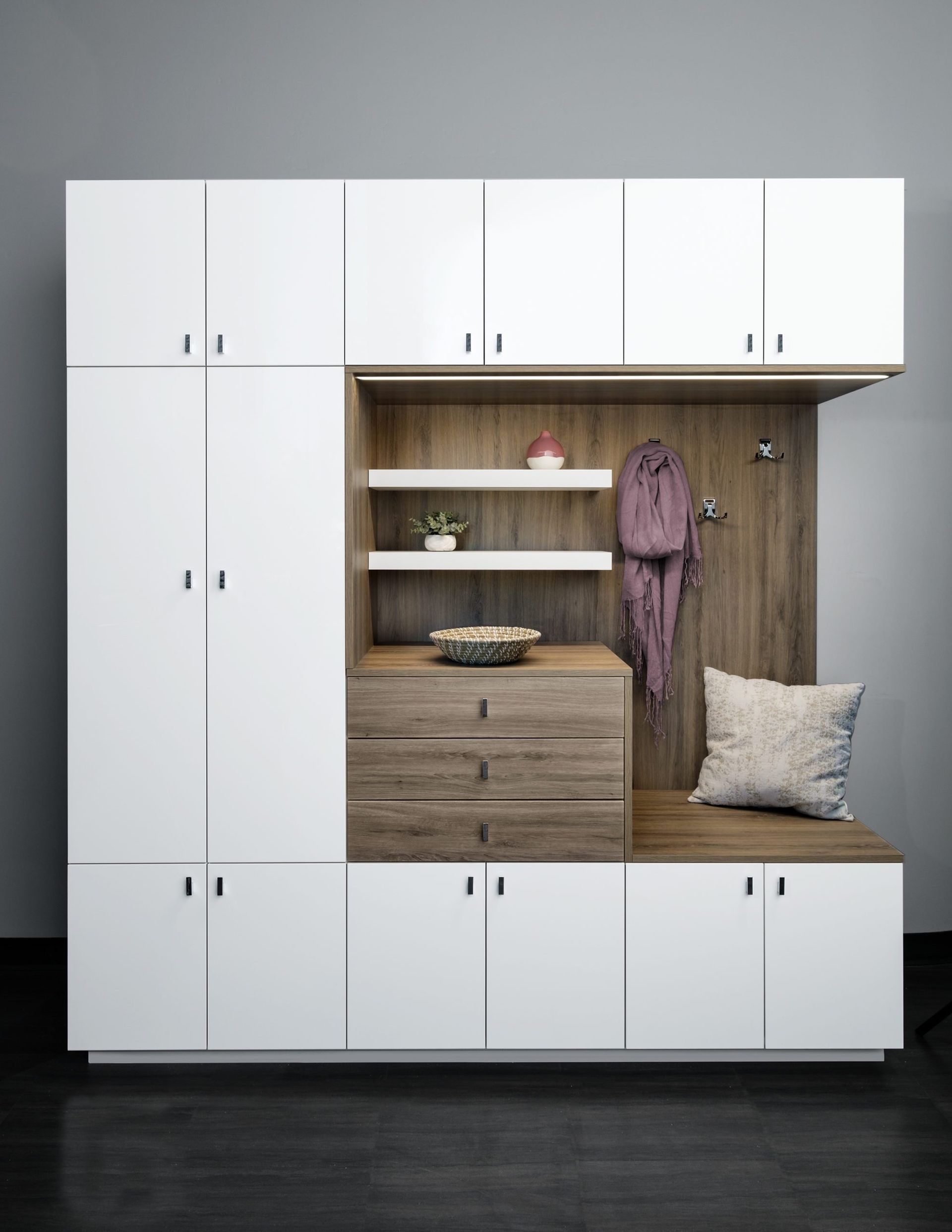 White modular wall cabinet with wood shelves, drawers, and a built-in bench nook with hooks and a pillow.