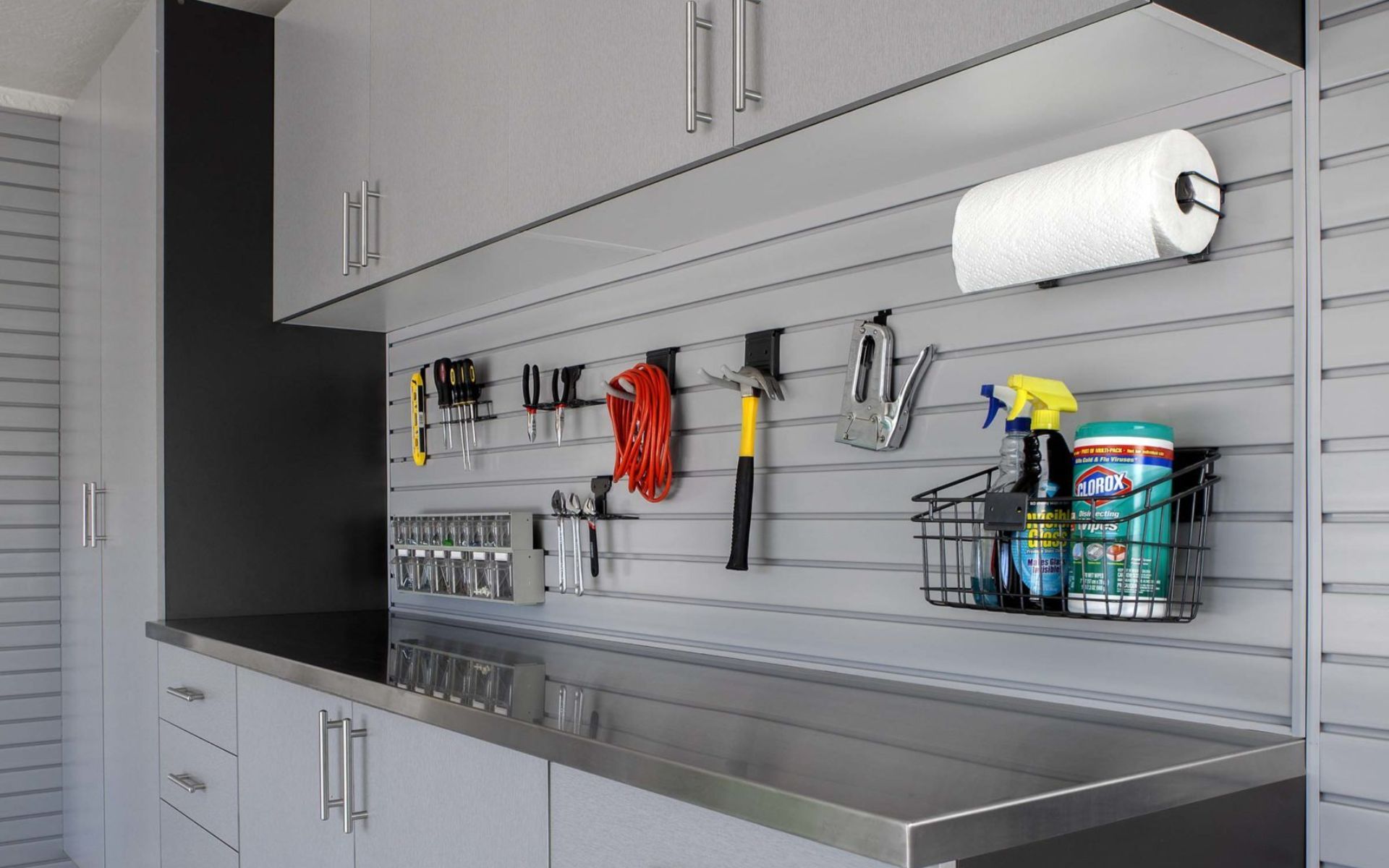 Wall-mounted garage tool organizer with hooks, shelves, and cleaning supplies above a metal workbench