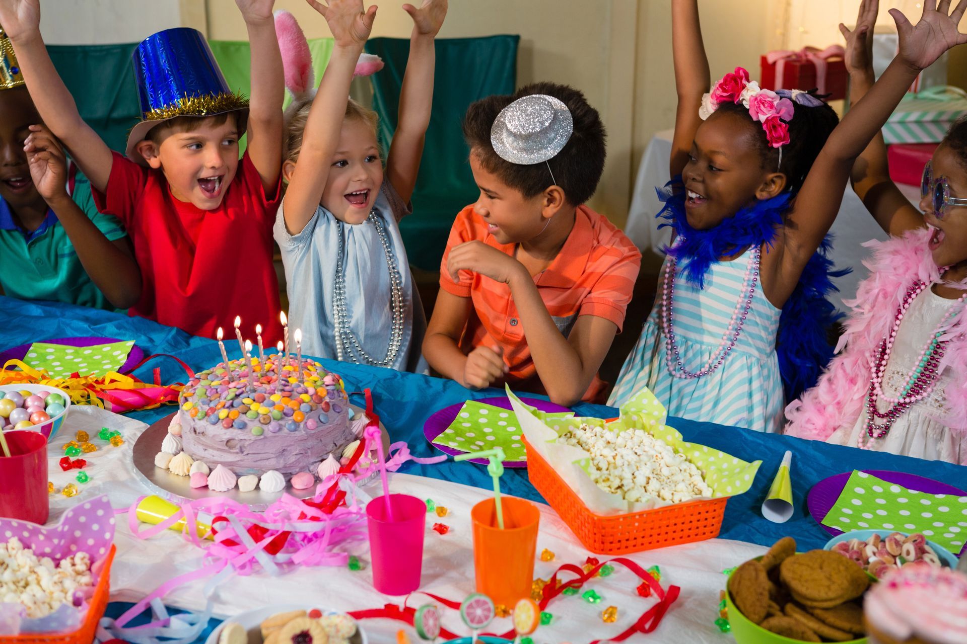 A group of cheerful children with arms raised over a table full of treats at a birthday party.