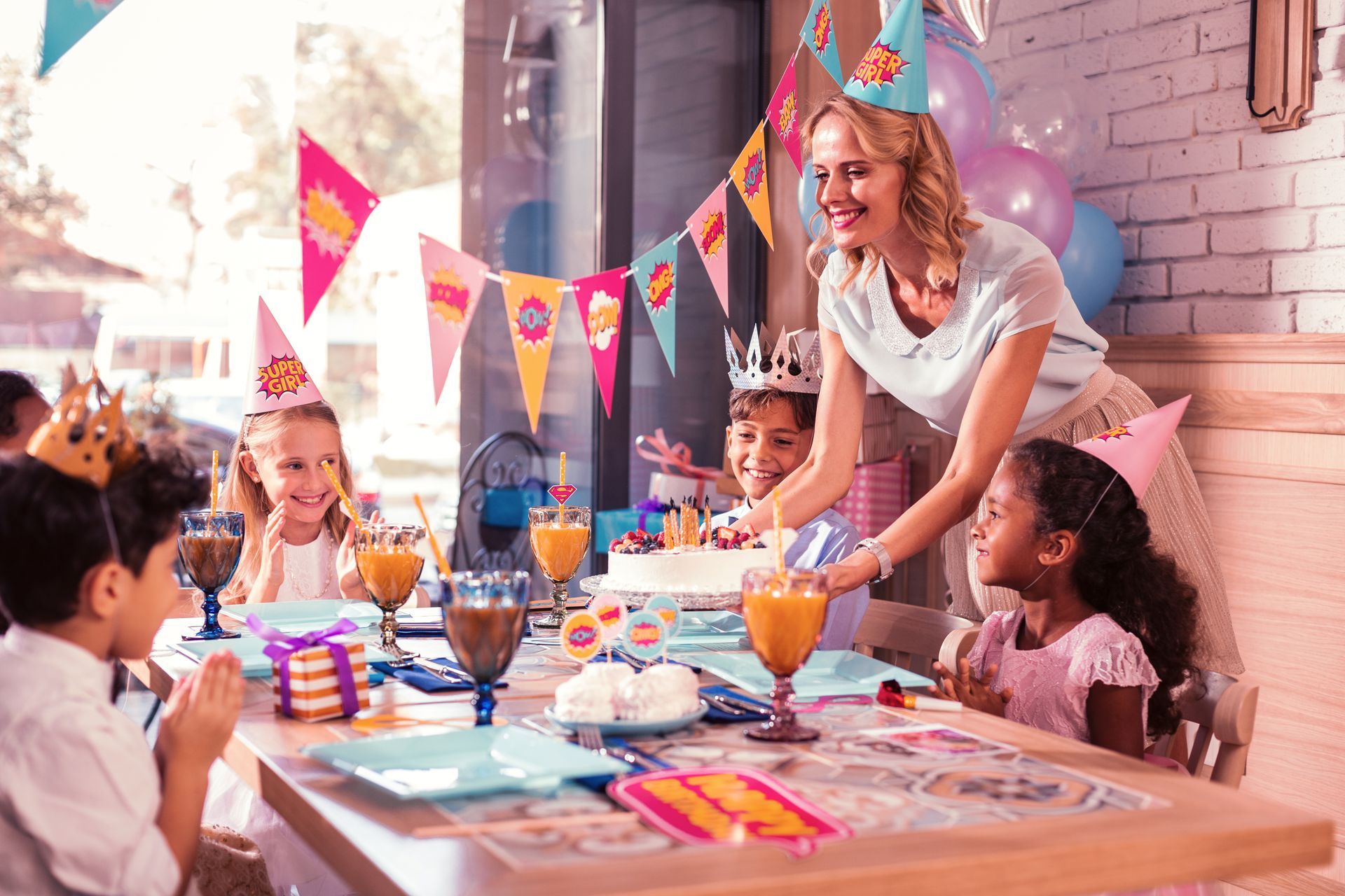 Adult placing a birthday cake on a table while children gather for a birthday celebration.
