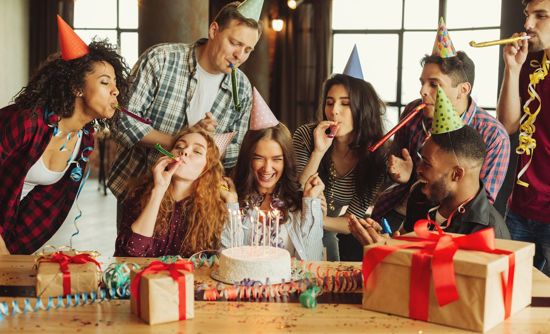 Group of people wearing party hats gathered around a birthday cake and wrapped gifts at a party. Group of people wearing party hats gathered around a birthday cake and wrapped gifts at a party.