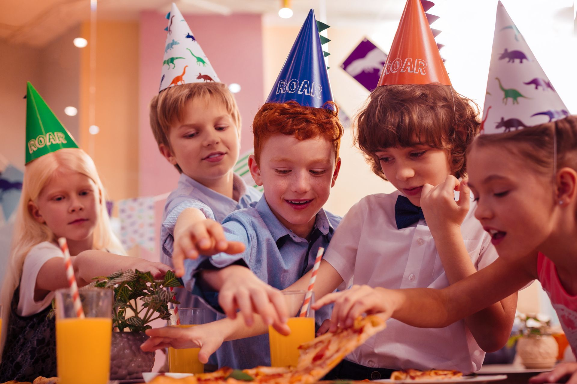 Kids in party hats reaching for pizza slices at birthday party.