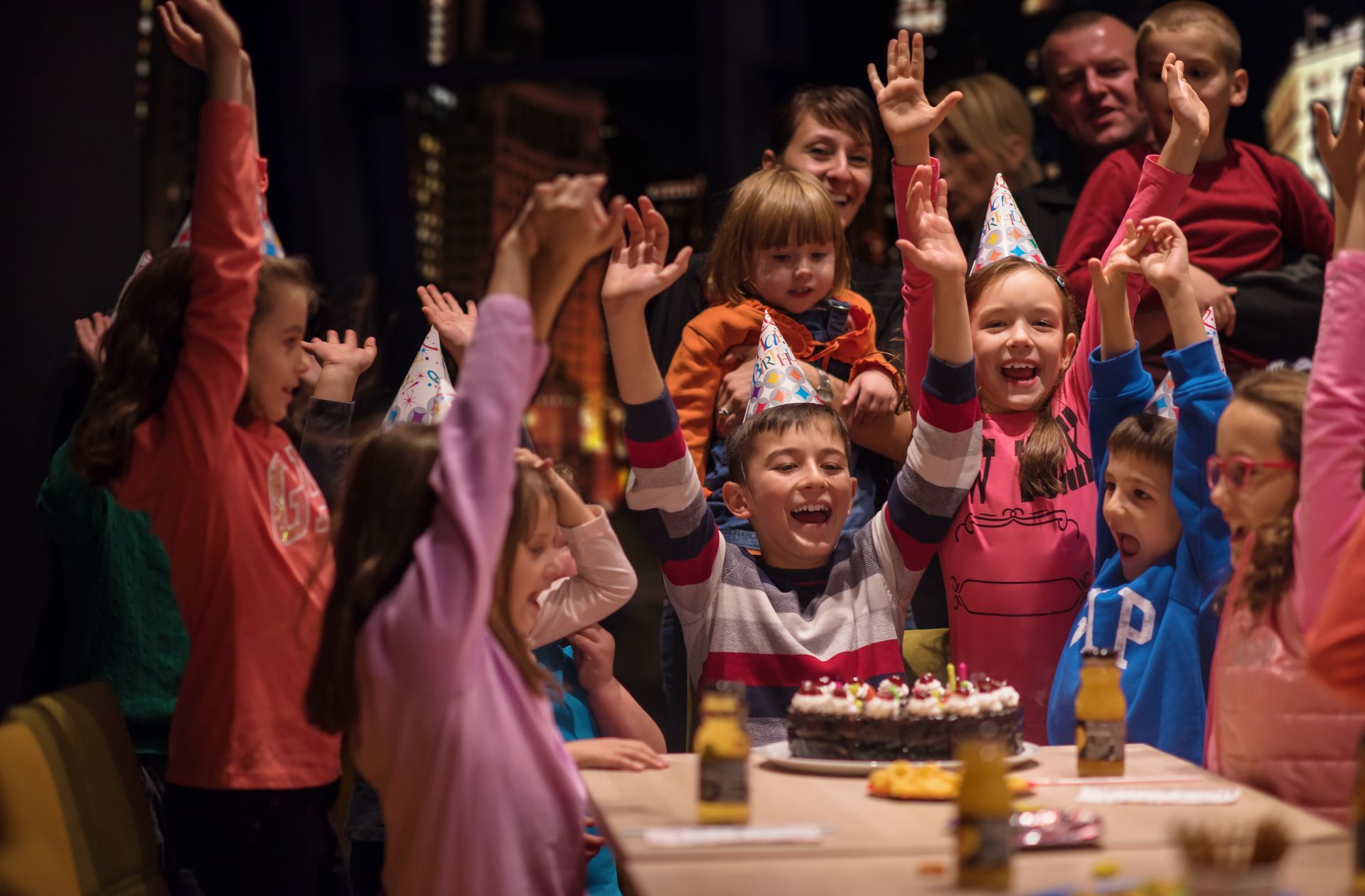 Group of children wearing party hats gathered around a birthday cake with adults.
