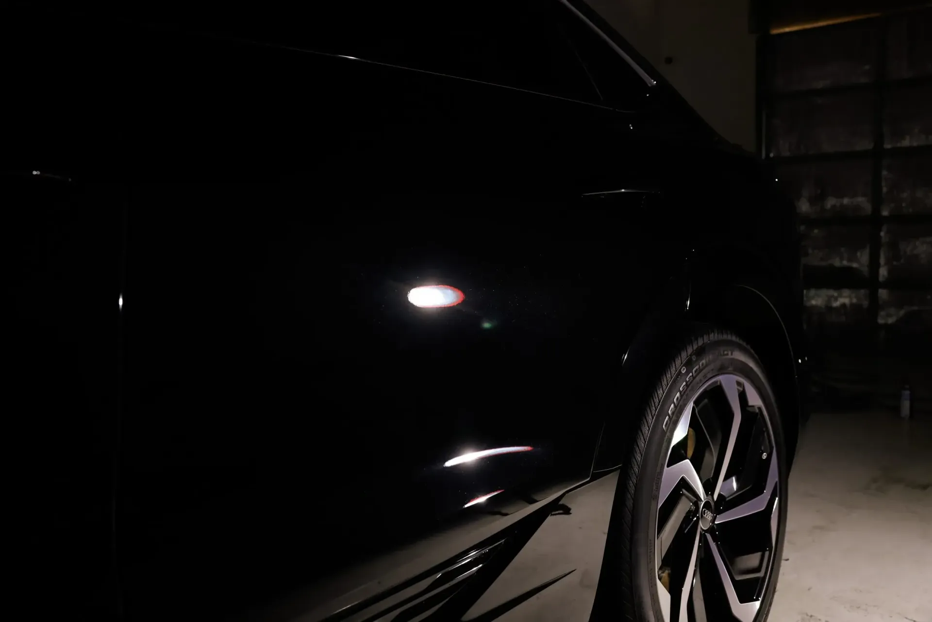 Black car side with a shiny rim, illuminated in a dark setting.
