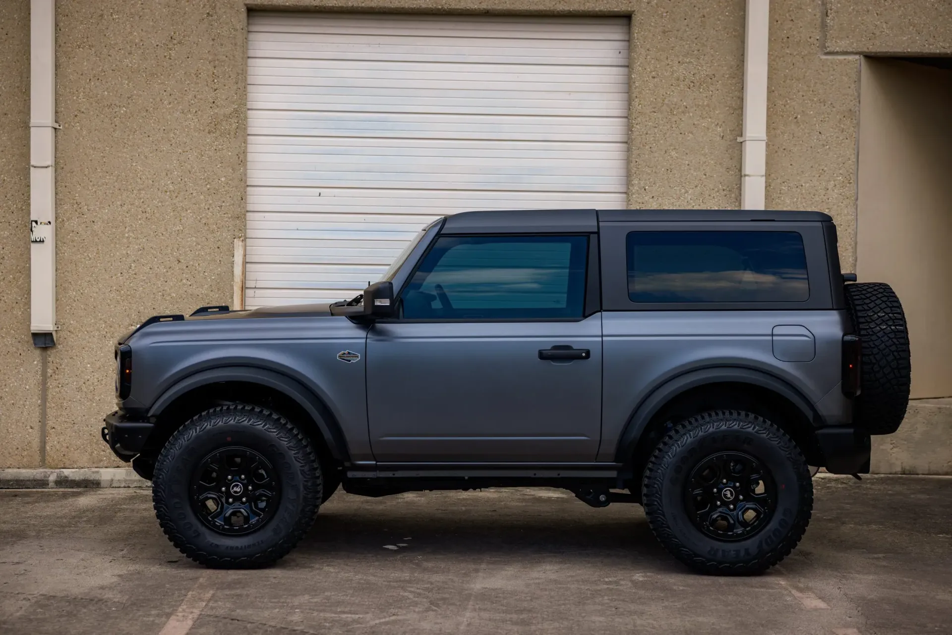 Gray Ford Bronco SUV with black wheels and a spare tire, parked in front of a tan building with a closed garage door.