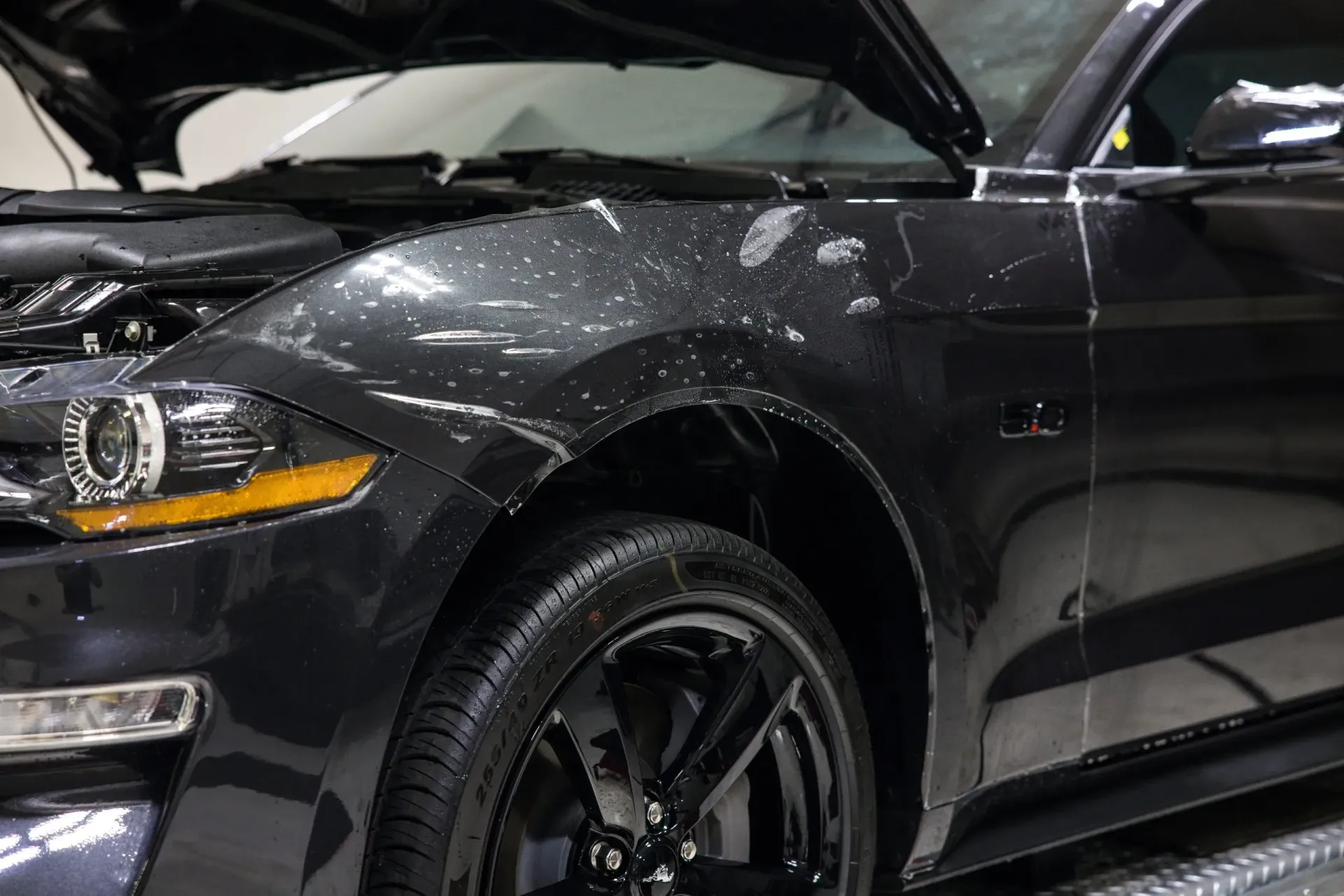 Black Ford Mustang with wet exterior; hood open, wheel and headlights visible.