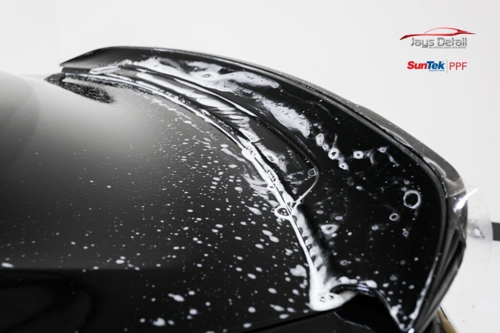 A car spoiler being covered with a clear protective film, with bubbles, in a shop setting.