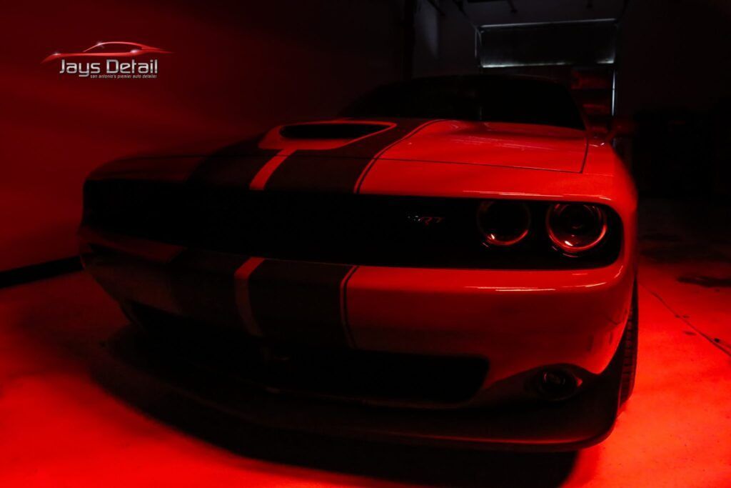 Red and black Dodge Challenger with racing stripe, lit with red light, inside a dark garage.