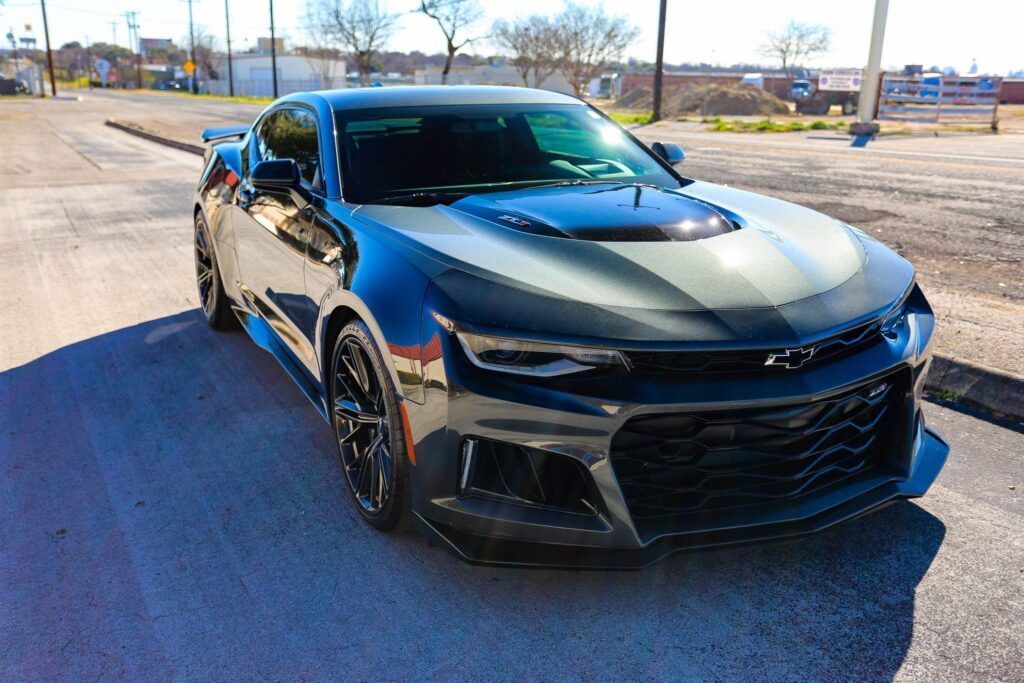 Dark gray Chevrolet Camaro sports car on a paved road, black wheels, gray stripes, sunny day.