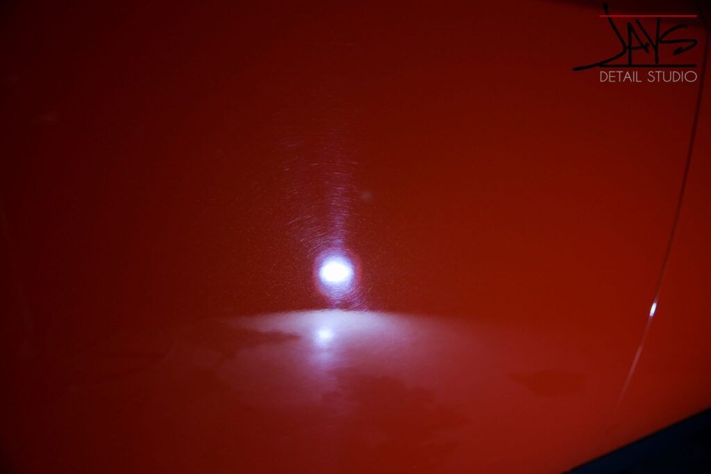 Close-up of red car paint with circular water spots and a bright reflection of a detailing light.