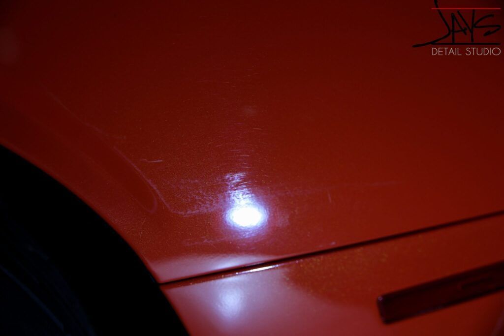 Close-up of red car paint with swirl marks and a light reflection, likely showing paint correction needed.