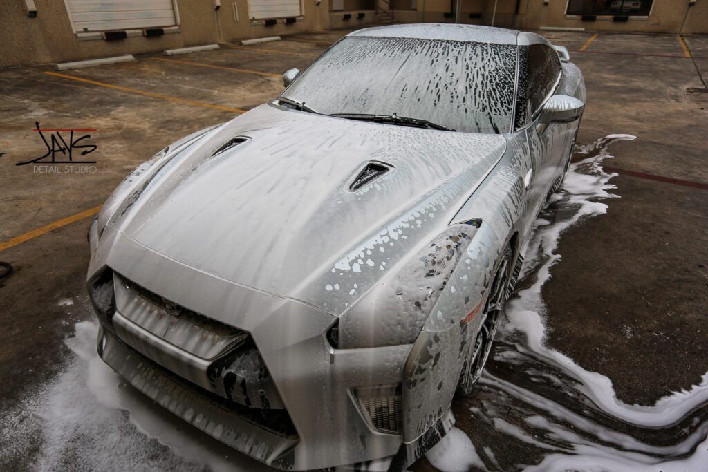 Silver Nissan GT-R covered in foamy soap suds, parked on a concrete lot, being washed.