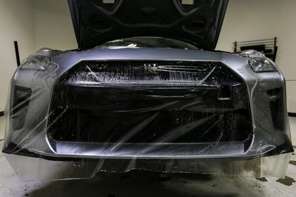A silver Nissan GT-R with protective film being applied to the front bumper, indoors.
