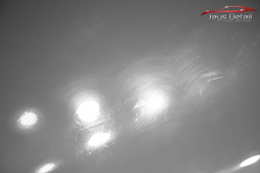 Gray car paint surface with swirling scratches visible under bright lights.