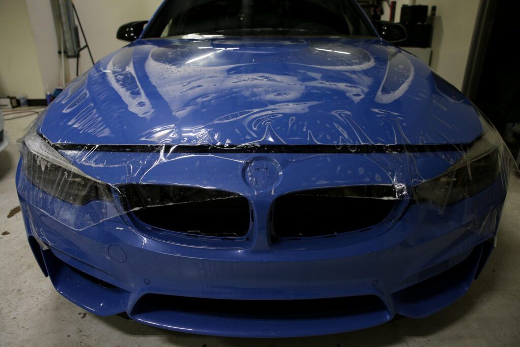 Blue car with protective film on the hood and headlights, likely being wrapped.