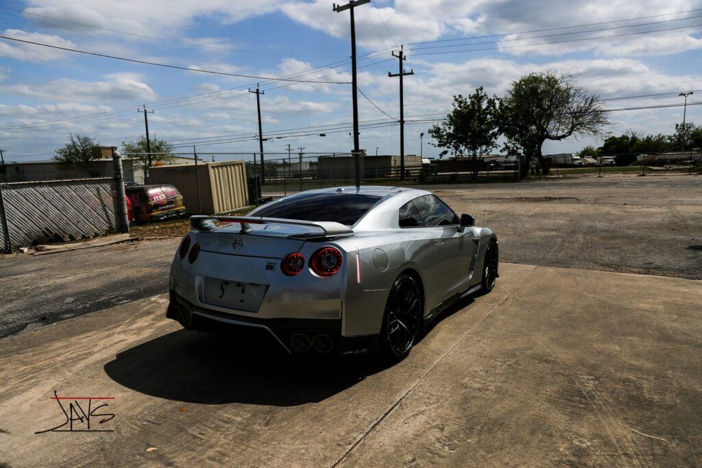 Silver Nissan GT-R sports car parked in a dusty lot near industrial buildings and power lines on a sunny day.