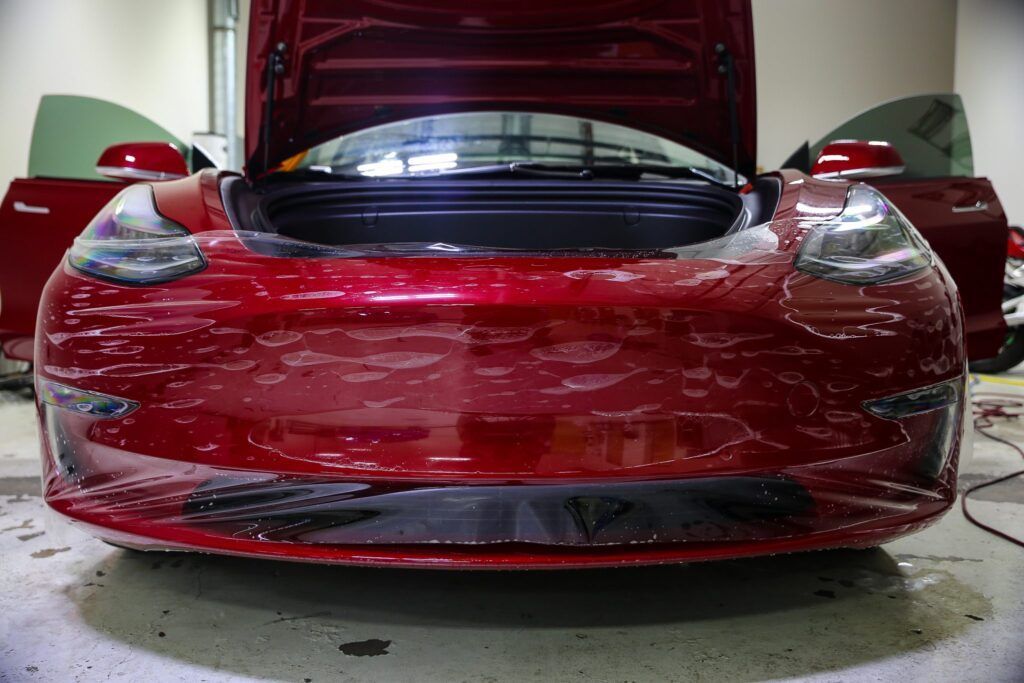 Red car bumper with protective film being applied in a garage.