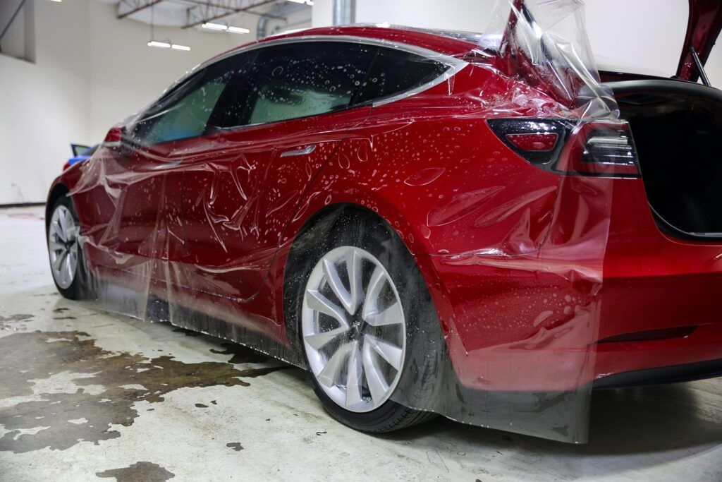 Red Tesla car being wrapped in protective film in a garage.
