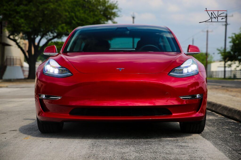 Red Tesla Model 3 electric car parked on a street, facing forward, with bright headlights on.