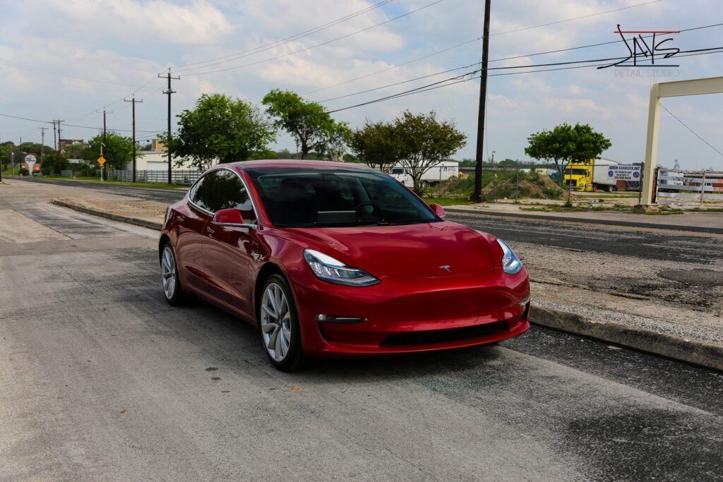 Red Tesla sedan on an asphalt road, parked near buildings and trees, on a cloudy day.