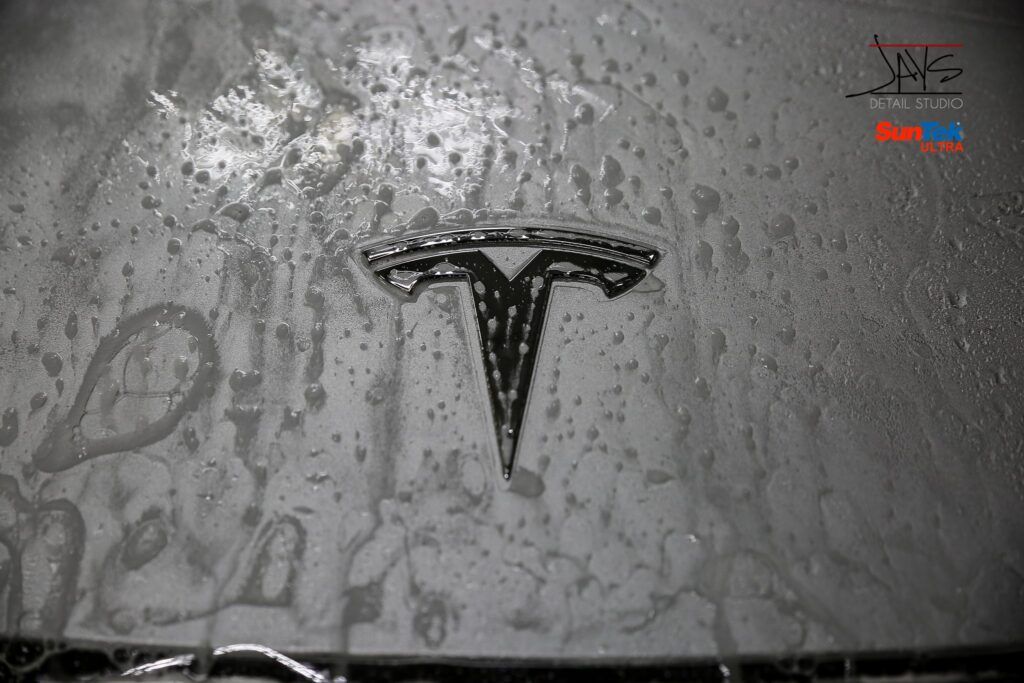 Tesla logo covered in soapy foam during car wash.