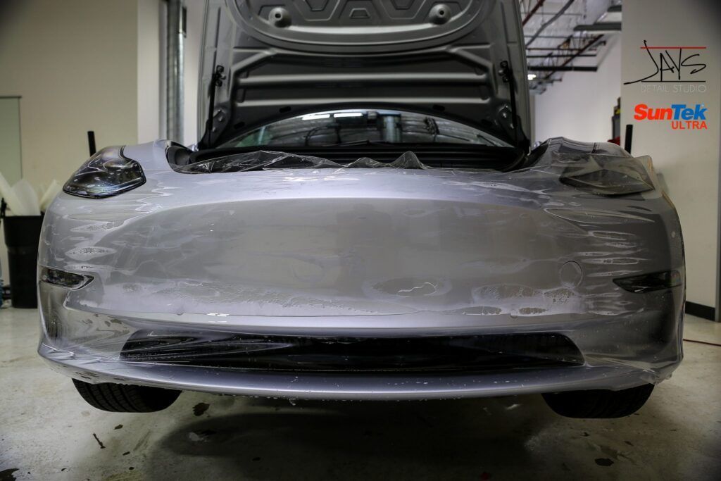 Gray car with open hood, covered in protective film in a garage.