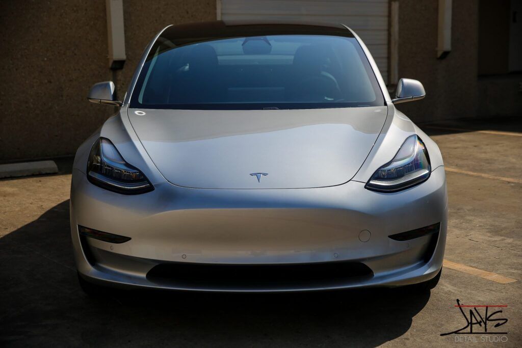 Silver Tesla Model 3 electric car parked in front of a building on a sunny day.