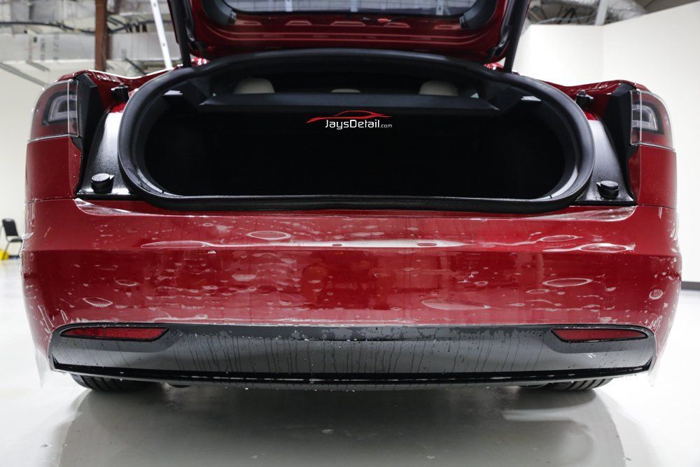 Red Tesla Model 3 rear with trunk open, showing cargo area. Tail lights and rear bumper visible.