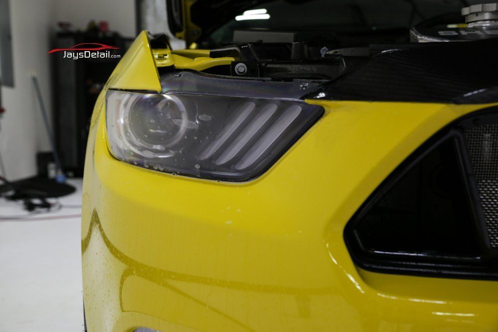 Yellow Ford Mustang's front end with a close-up of the headlight, under an open hood, in a shop.