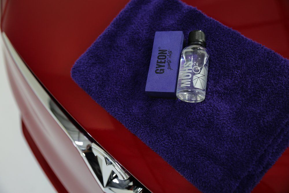 A red car hood with detailing supplies: purple towel, applicator, and ceramic coating.