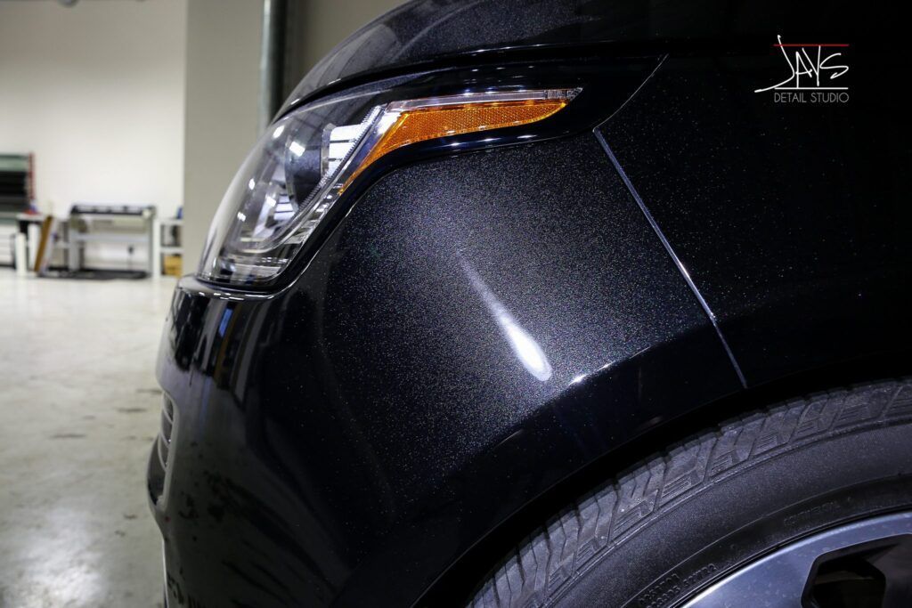 Black car's front with a textured finish, showing headlight, tire, and reflective highlights.
