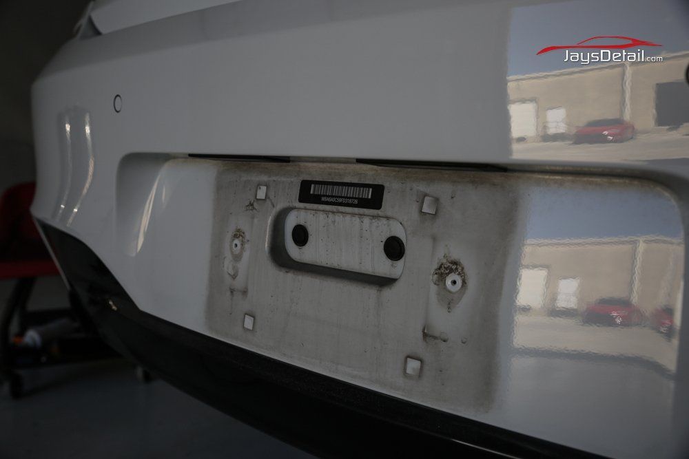 Close-up of a dirty car bumper. A license plate mount is visible, showing grime and dirt. Reflective surface.