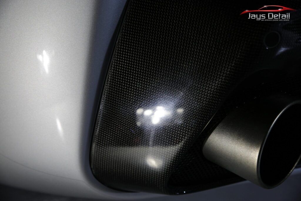 Close-up of a car's carbon fiber panel near a chrome exhaust pipe, reflecting bright lights.